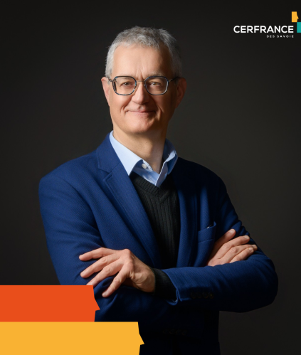 Portrait d'un homme à lunettes, bras croisés, portant une veste bleue sur un fond sombre.