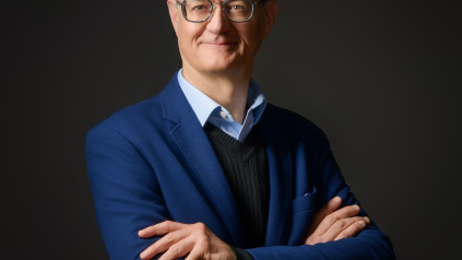 Portrait d'un homme à lunettes, bras croisés, portant une veste bleue sur un fond sombre.