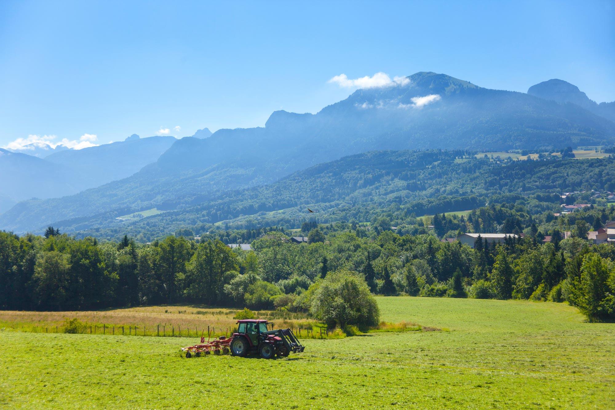 Un tracteur rouge équipé d'une faneuse travaille dans un grand champ vert, avec une vue panoramique sur des montagnes boisées et un ciel bleu clair en arrière-plan