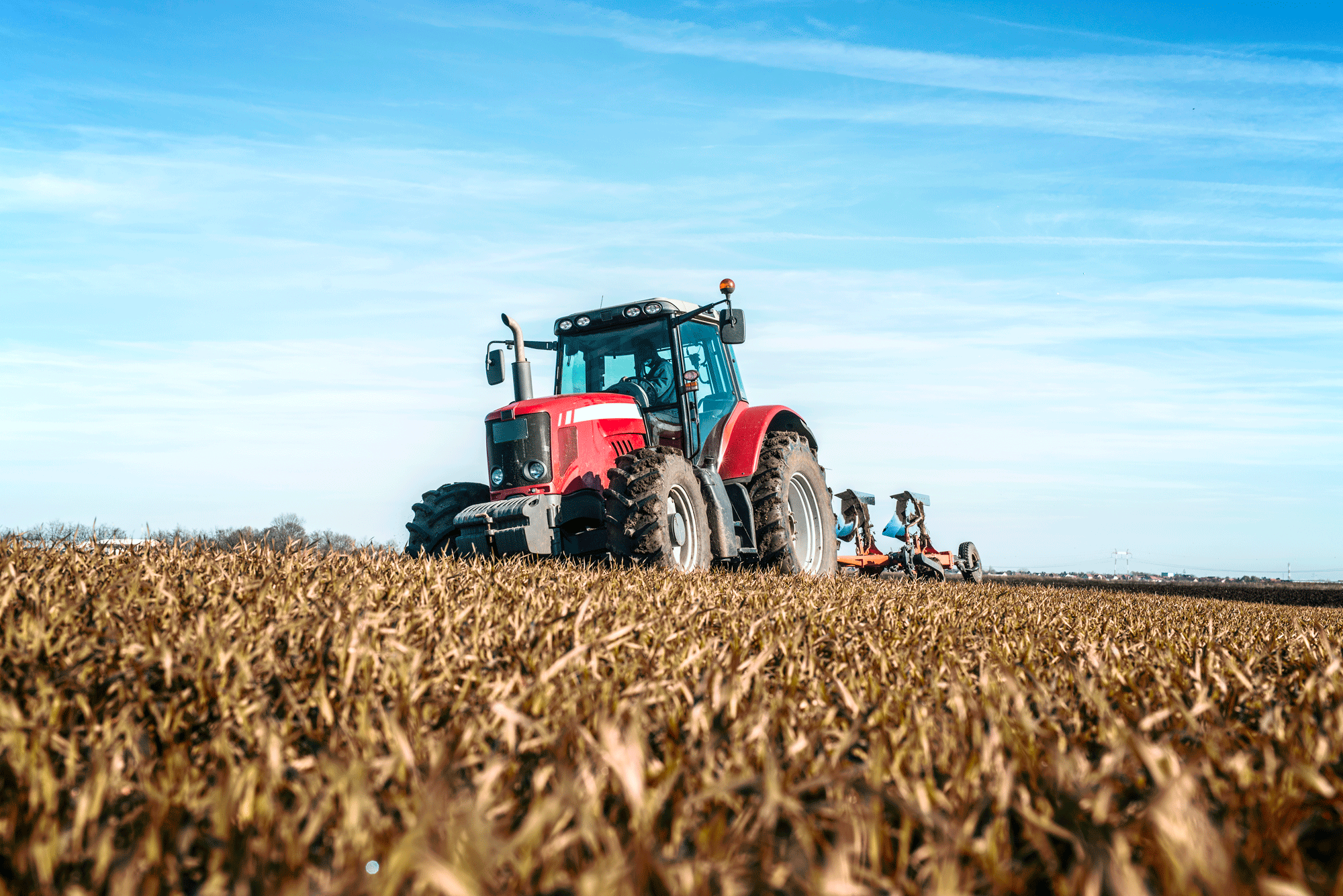 Un tracteur agricole rouge moderne équipé d'une charrue travaille dans un vaste champ sous un ciel bleu dégagé.