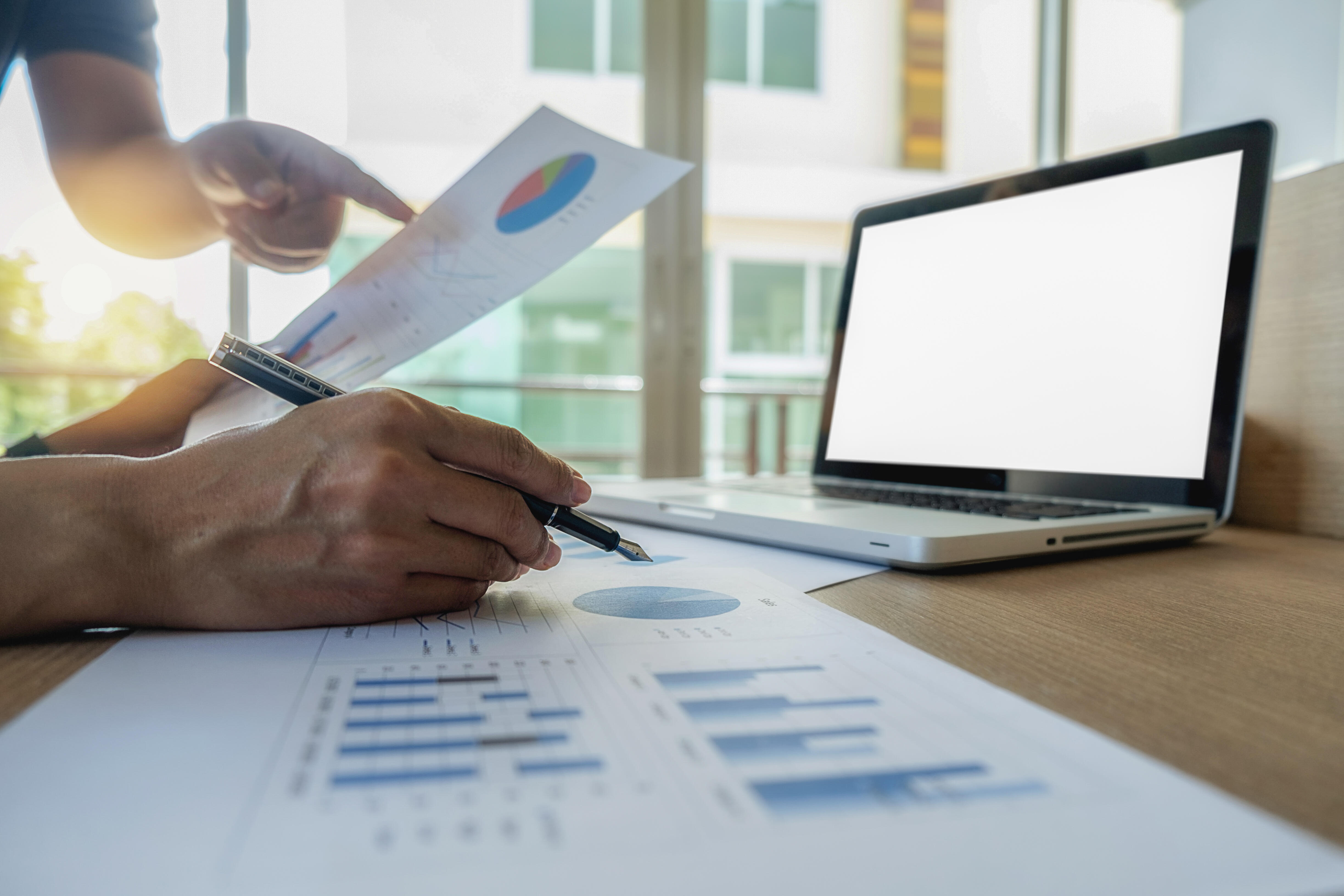 Deux professionnels analysent des documents financiers et des graphiques devant un ordinateur portable, dans un environnement de travail lumineux.
